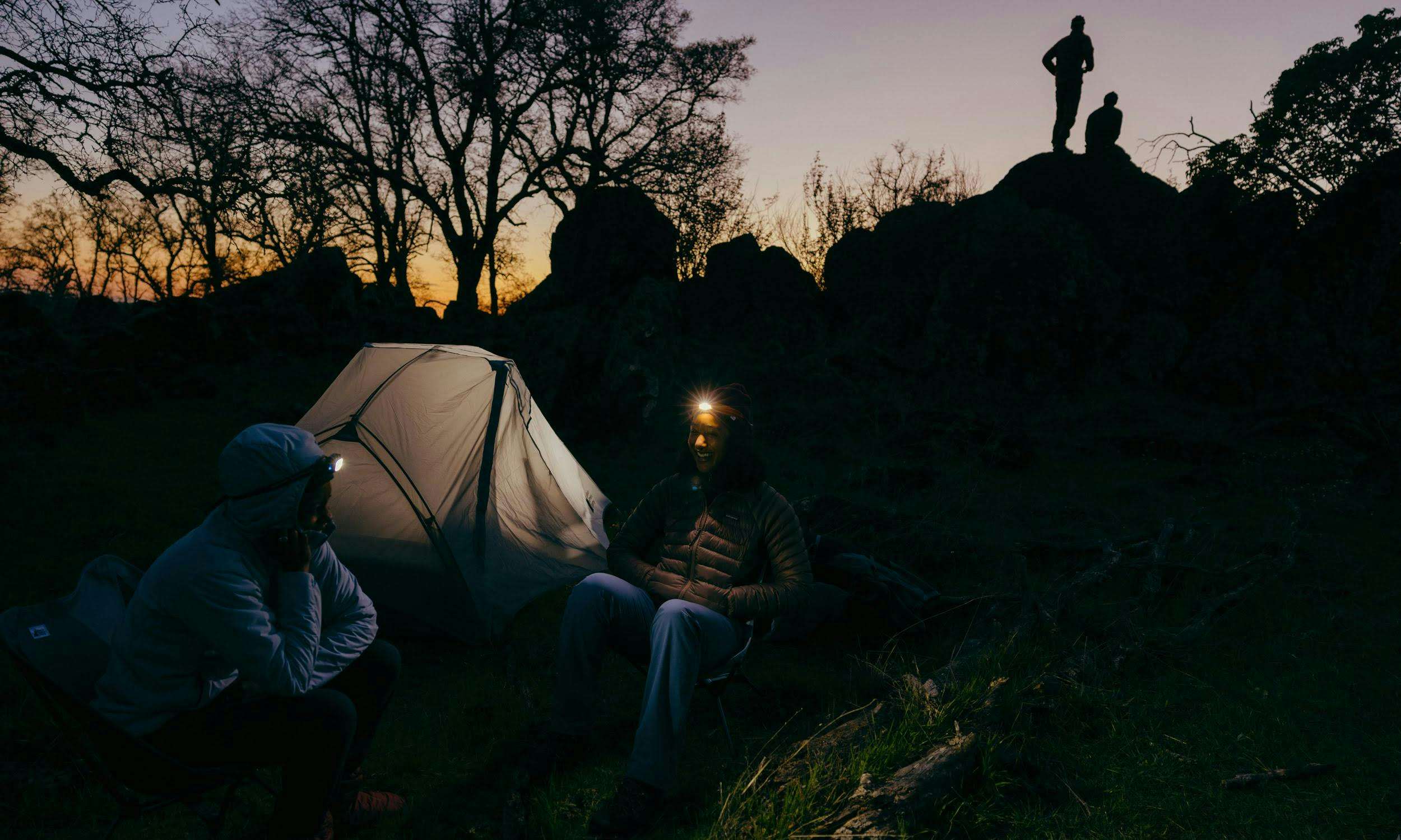 Four people camping outside without a campfire, using headlamps for light. 