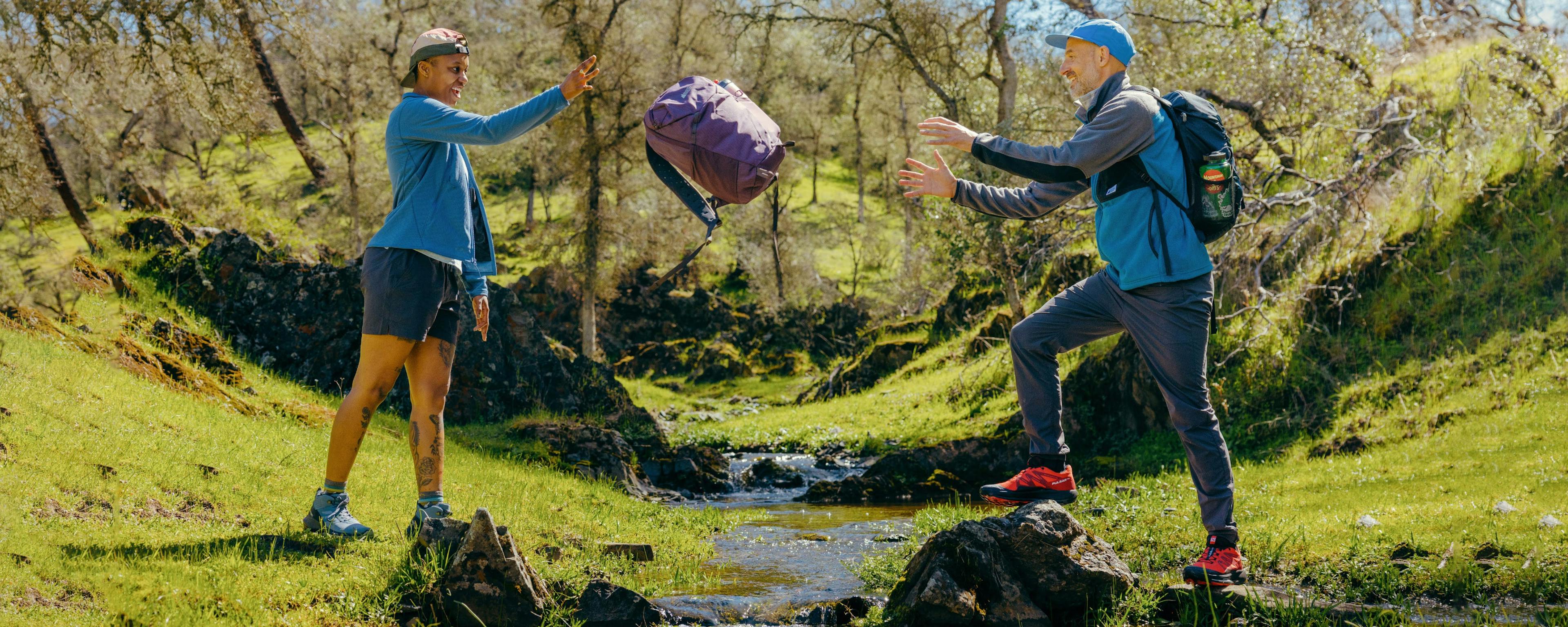 Two hikers crossing a small stream; one hike is tossing a backpack to the other