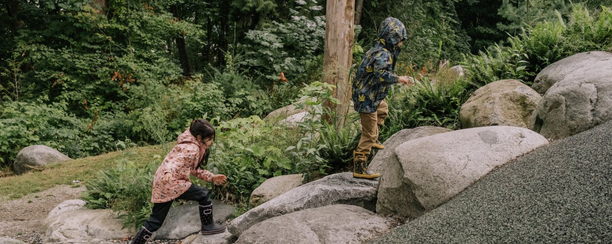 Two kids walking up large boulder path in forested area wearing rain jackets and rain boots