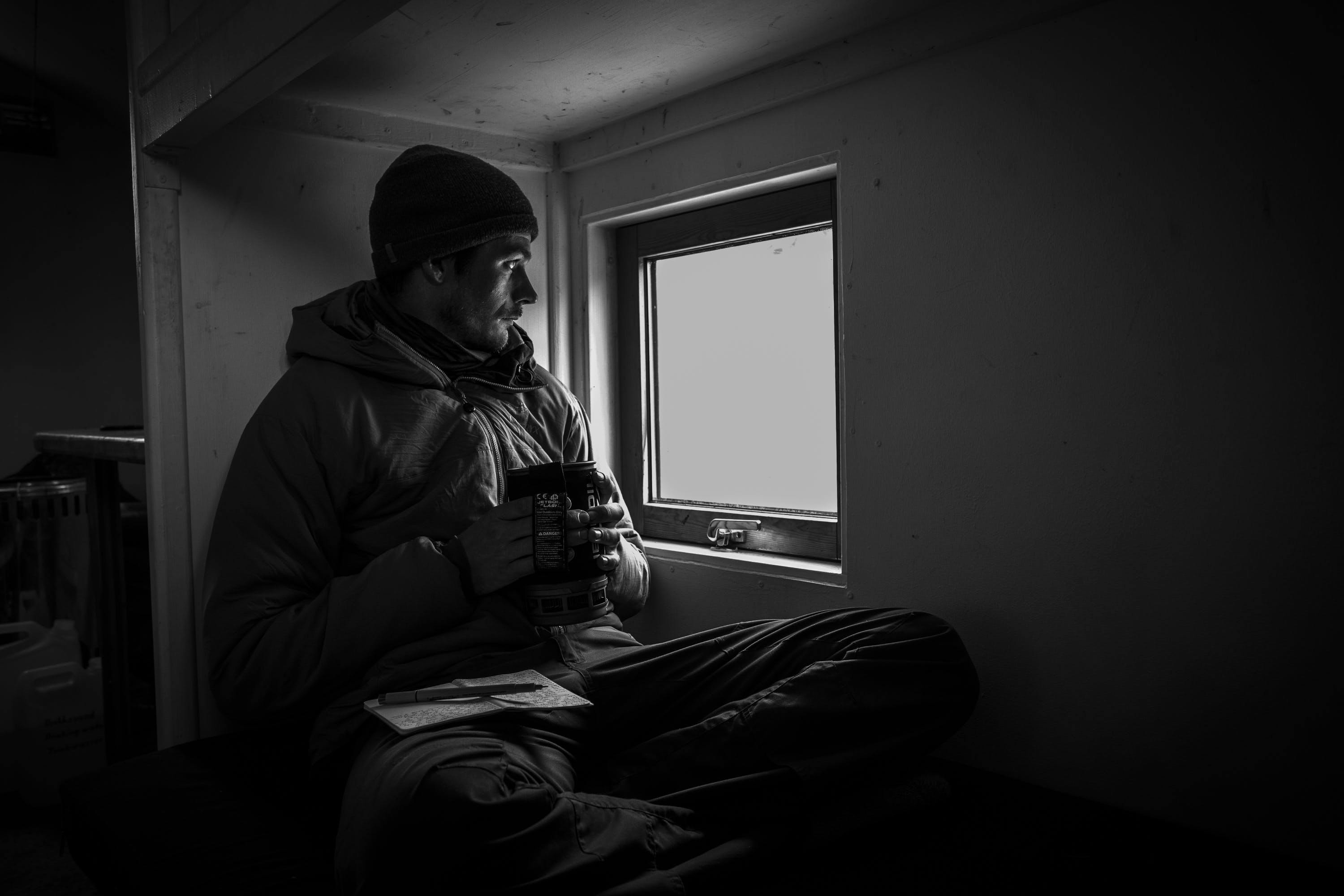 A man holds a mug of tea and looks out the window of a dark hut.
