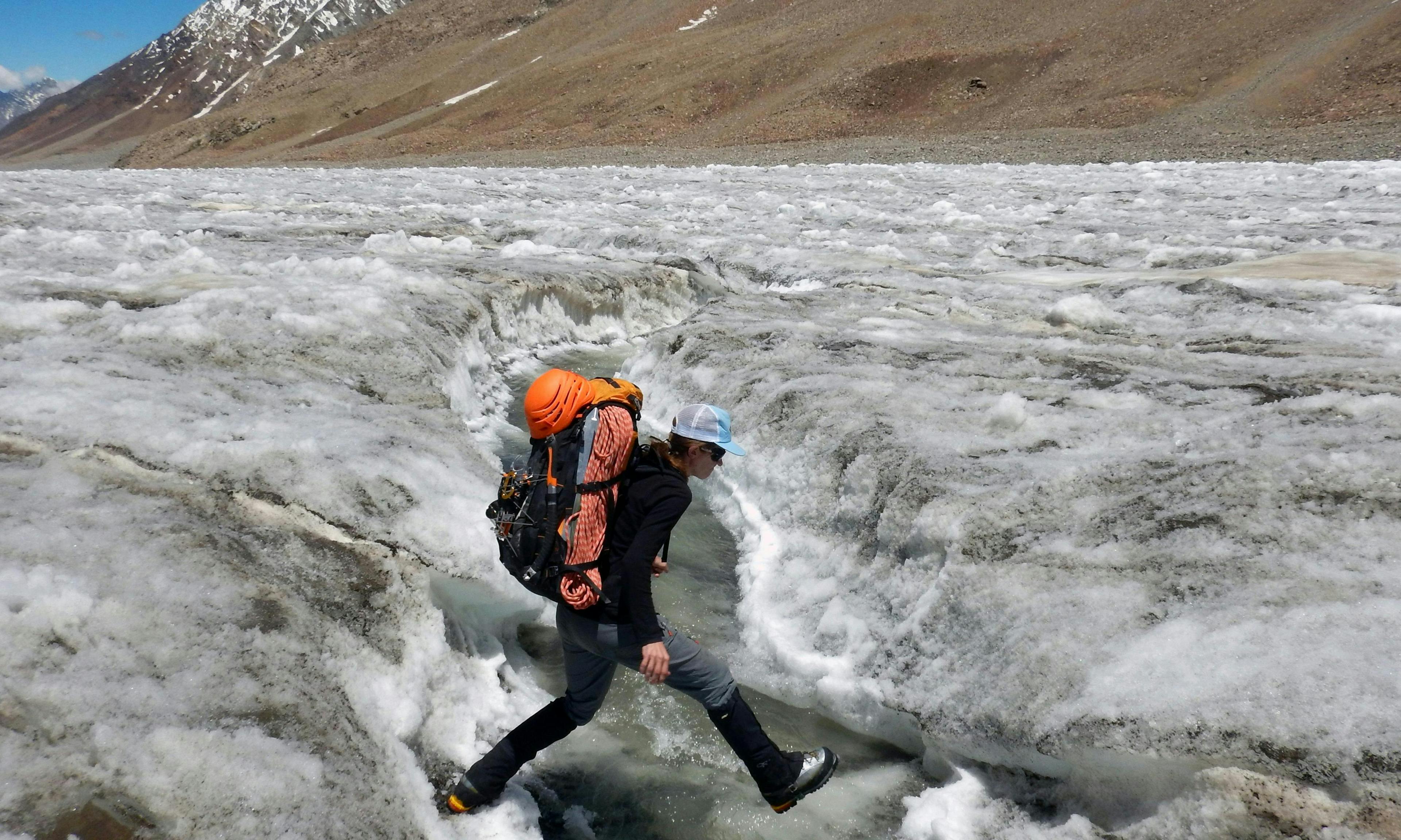 A person wearing a backpack and mountaineering gear hops over a crevasse on a glacier.