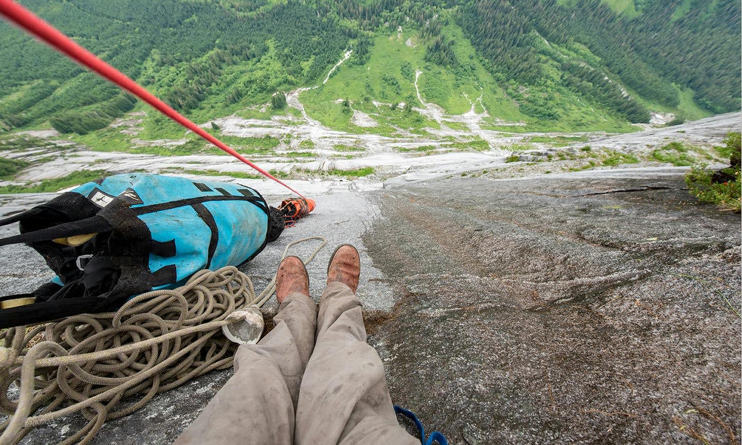 A person's feet and backpack on the edge of a rock wall, looking down at the green valley below.