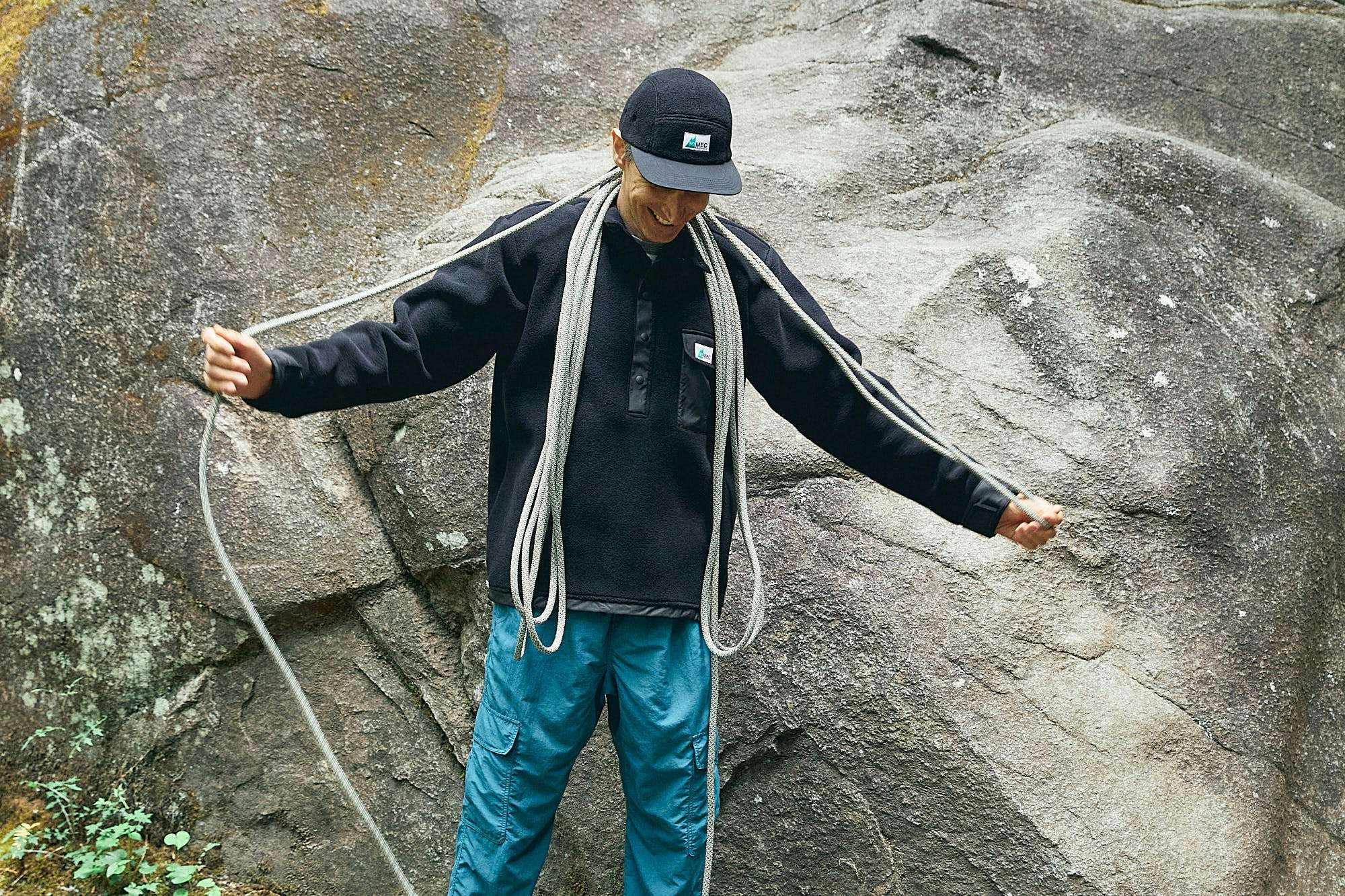 Person wearing a black MEC cap in front of a boulder coiling climbing rope