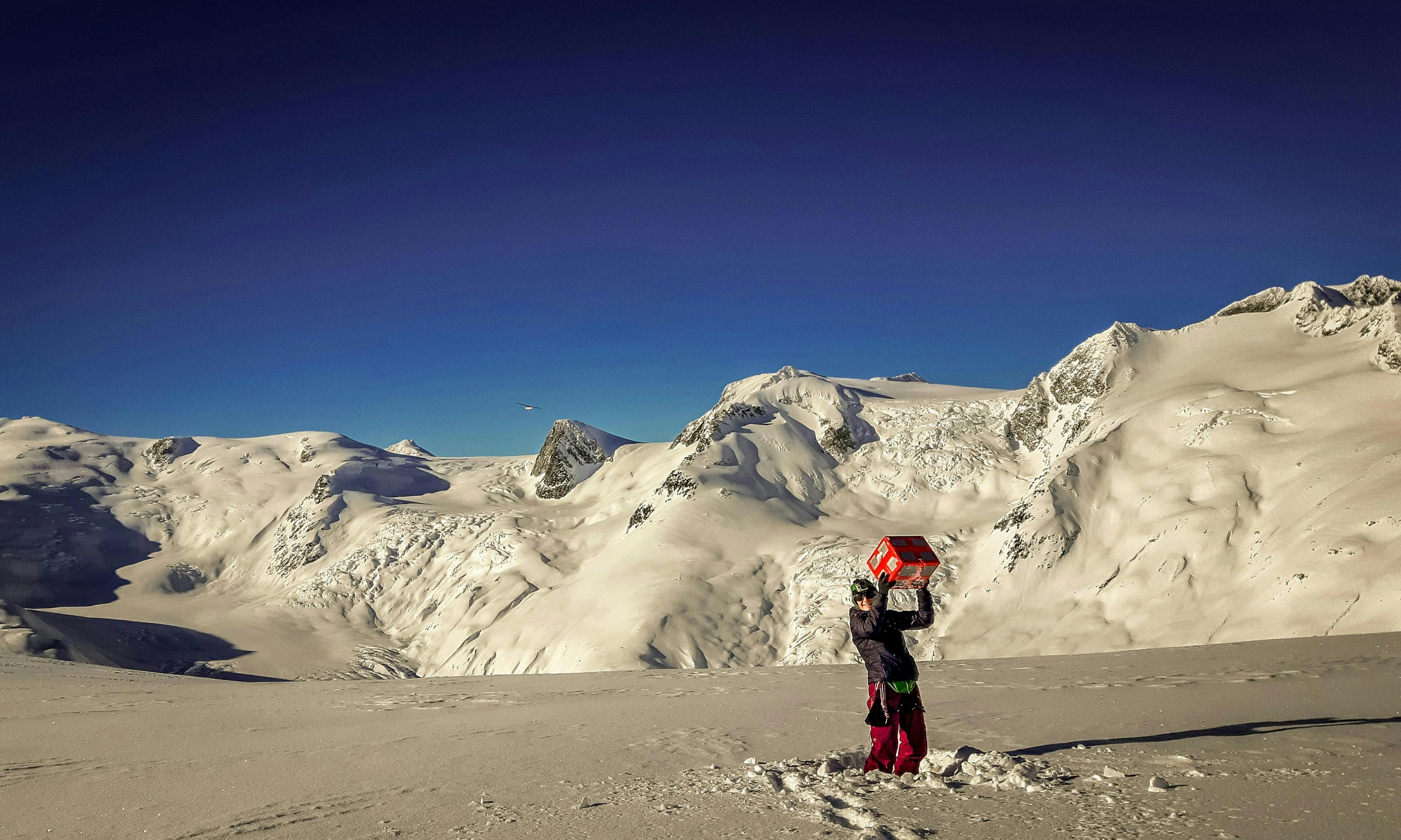 A woman holds a food box aloft while standing in the middle of a snowfield, surrounded by mountains.