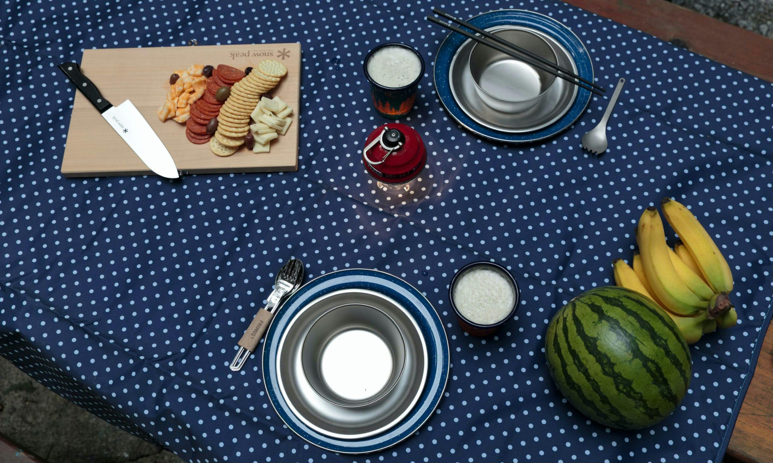 Picnic set-up with camping utensils, bananas, watermelon and a board with crackers and cheese