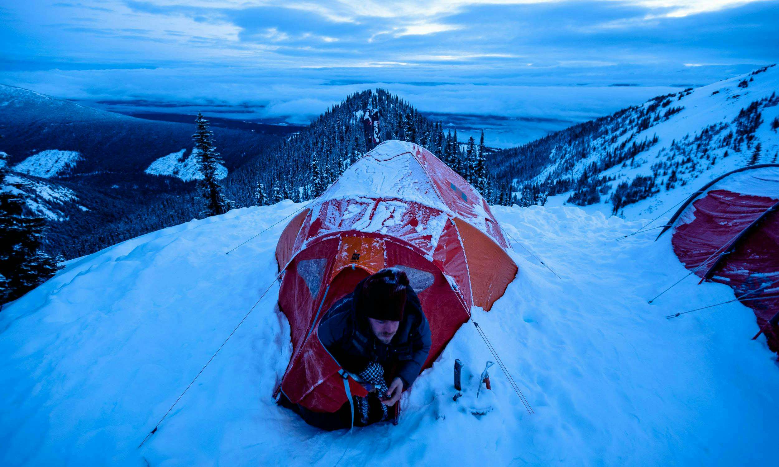 A man leans out of an orange tent perched on a snowy mountain, with a view spread behind him.
