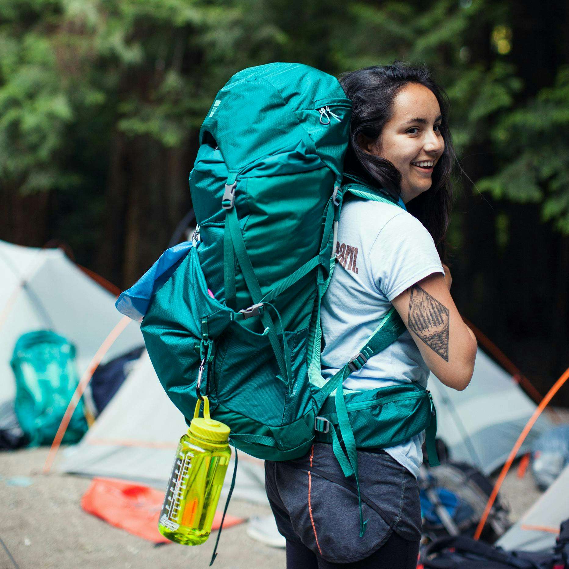 A young woman wearing a hiking backpack looks back over her shoulder and smiles.