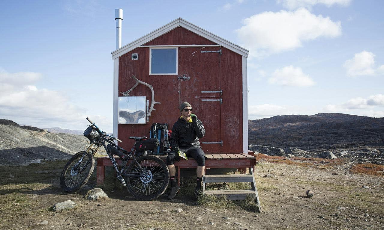 A man and his mountain bike sit outside a small red house in a remote area.