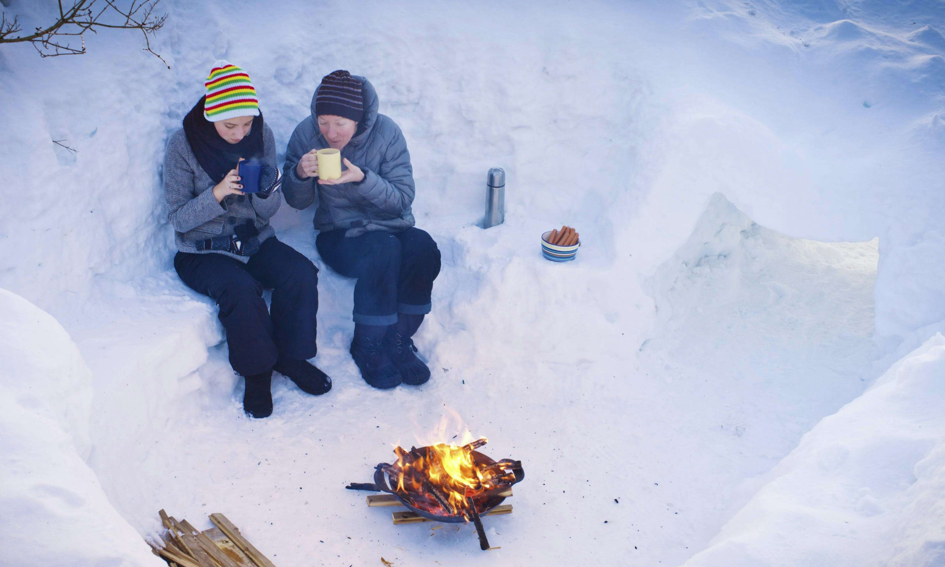 Two people sitting in the snow, sipping hot drinks with a Thermos nearby