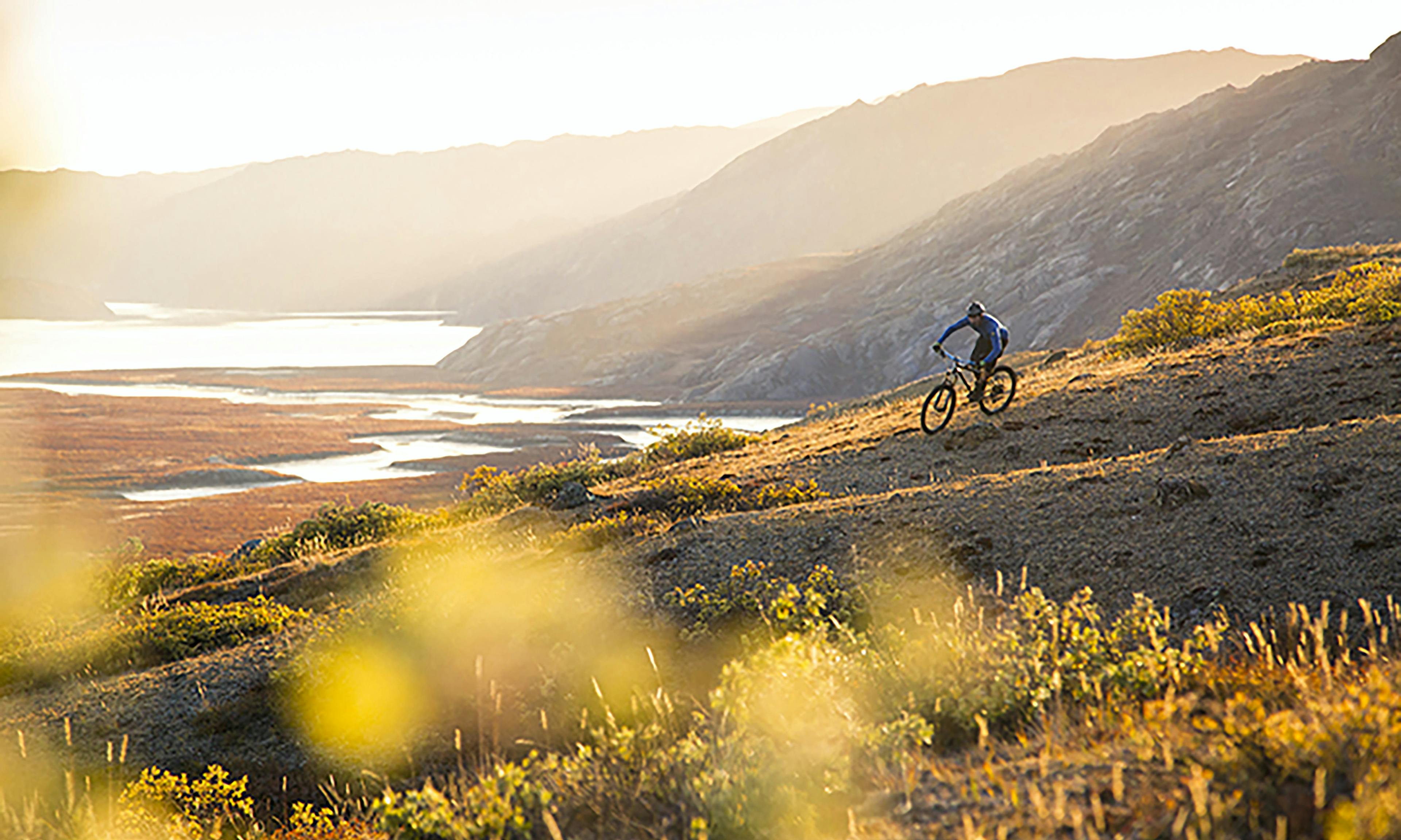 A man rides a bike along a grassy ridge at sunset, with mountains in the background.