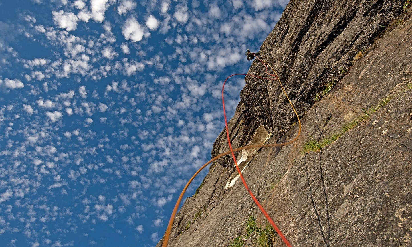 A climber far above the viewpoint shakes the orange ropes so they wave away from the rock face, silhouetted against a blue sky dotted with clouds.