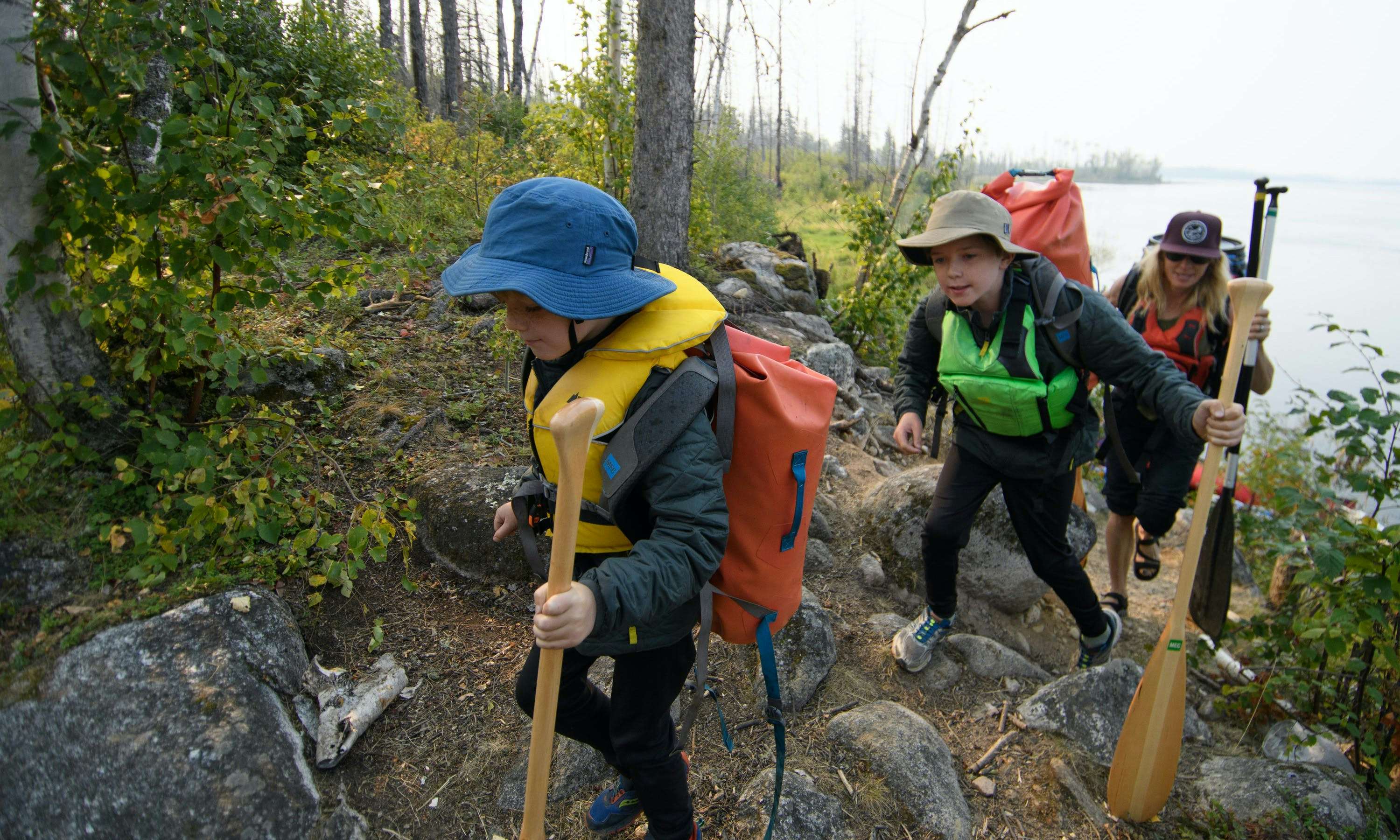 Two boys and their mother hike up a rocky trail carrying their canoe paddles and camping gear.