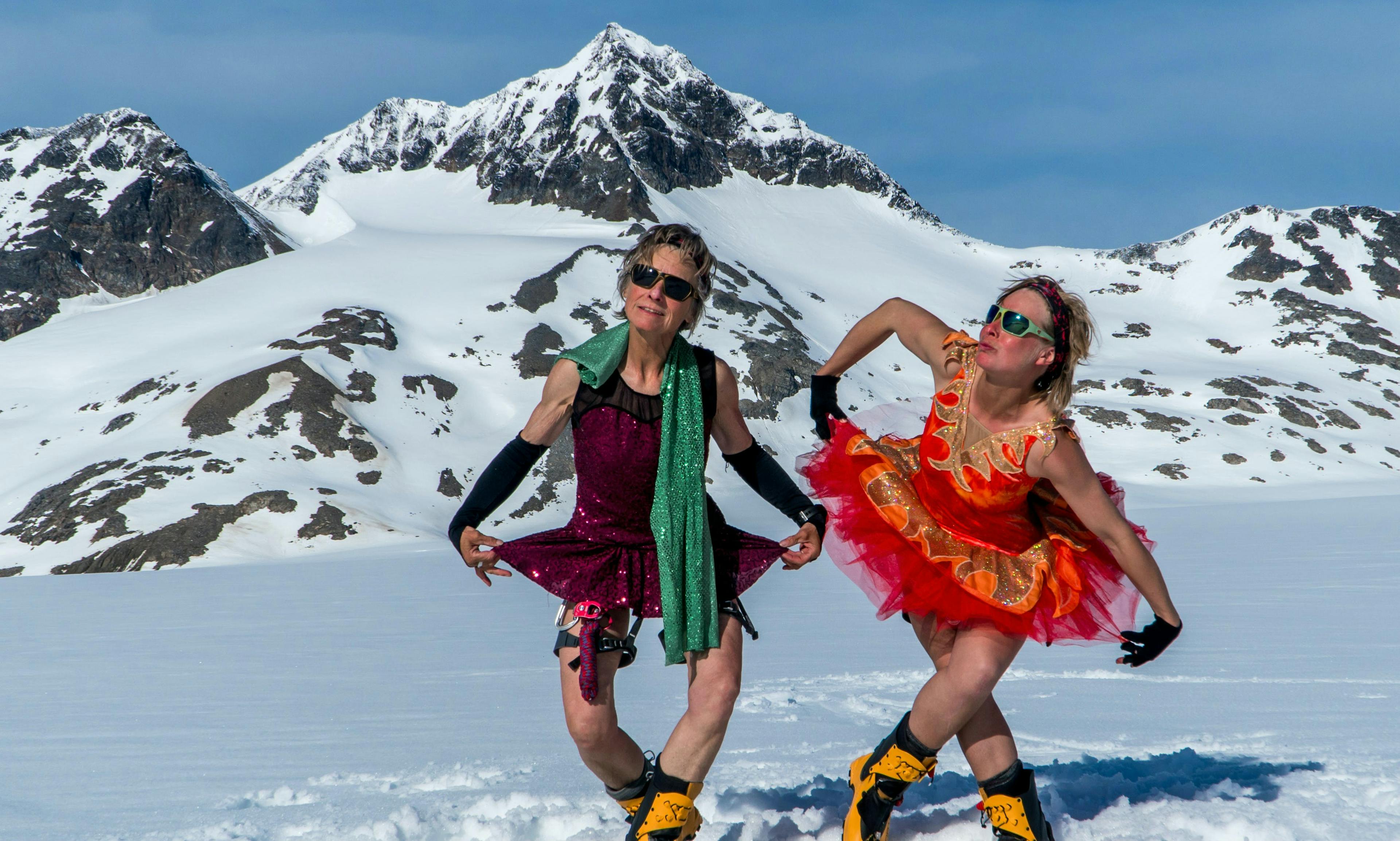 Two women curtsey while wearing shiny, flamboyant tutus in the middle of an alpine snowfield.