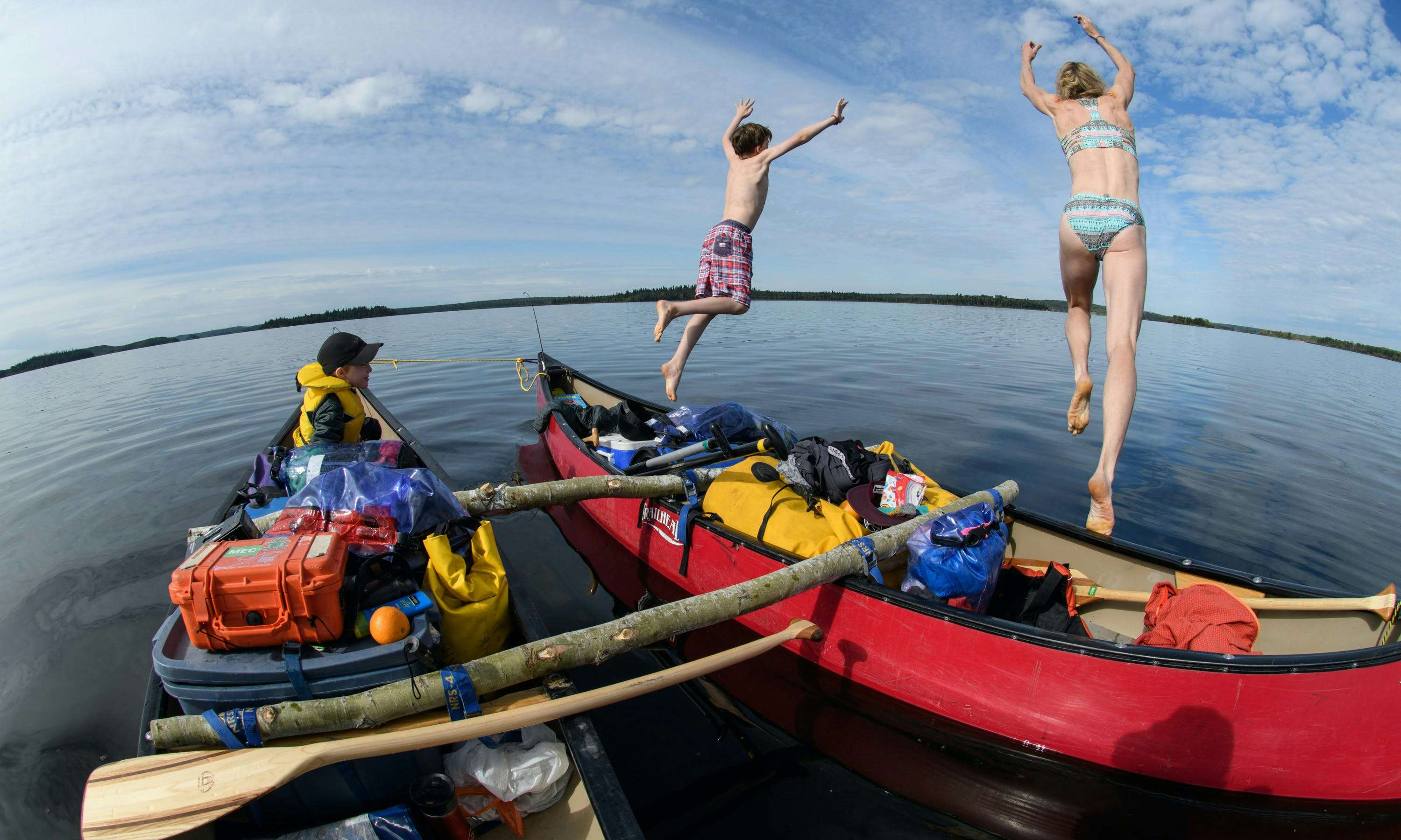 A woman and a boy leap out of their canoe into the water as another boy watches from a second canoe.
