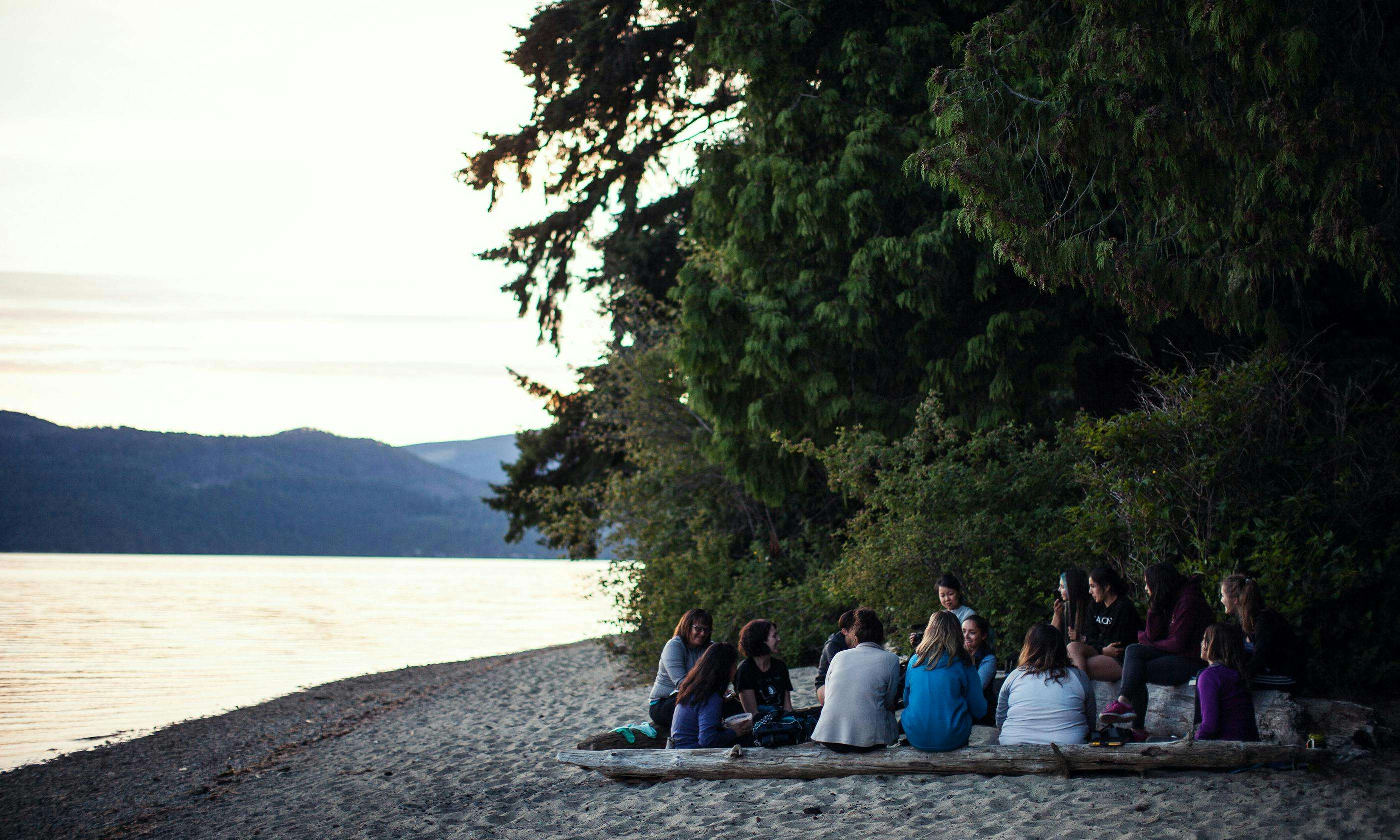 A group of young women sits in a circle on a beach as the sun sets.