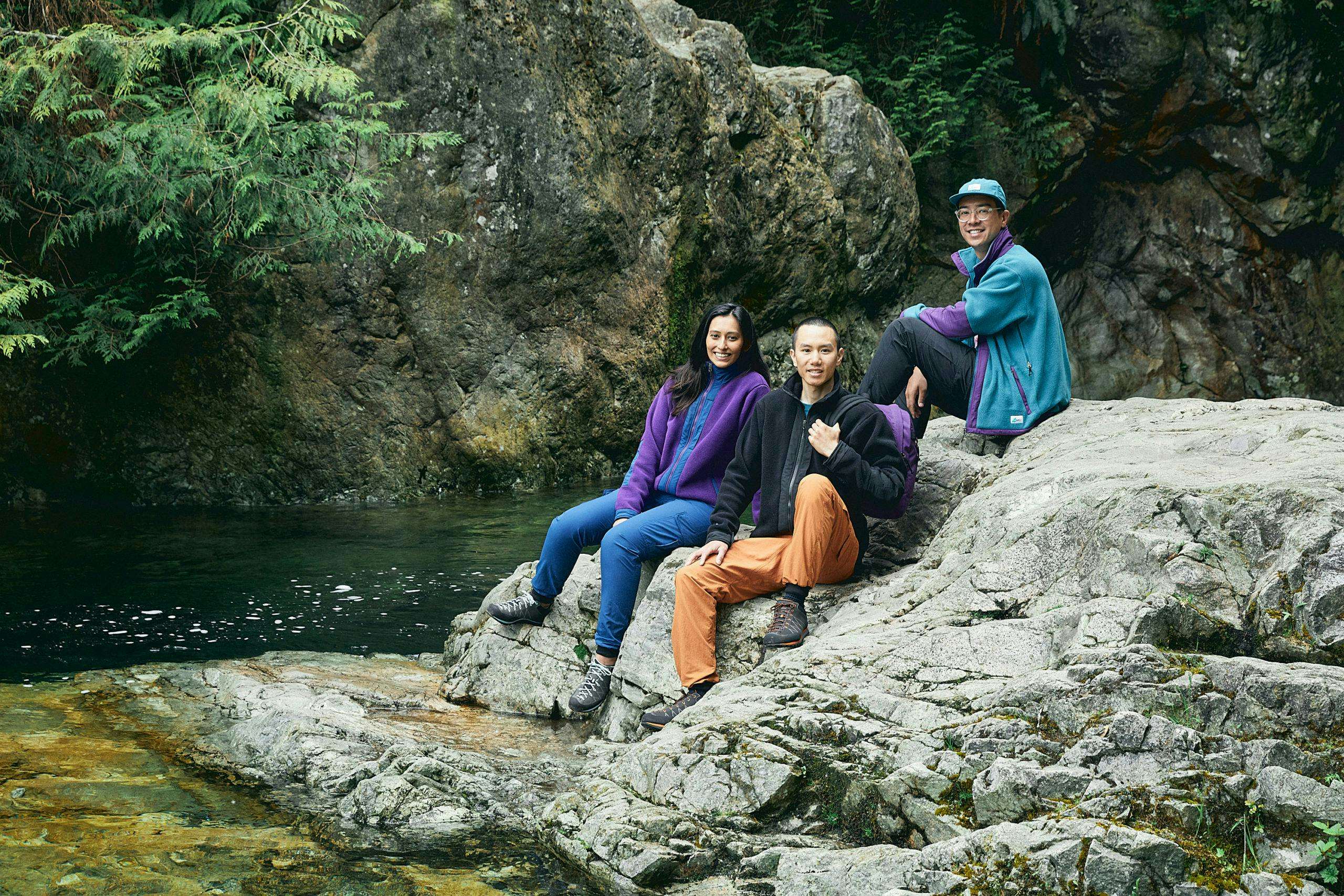 Three people sitting on rocks next to a river, wearing MEC fleece jackets that look like they're from the 1990s