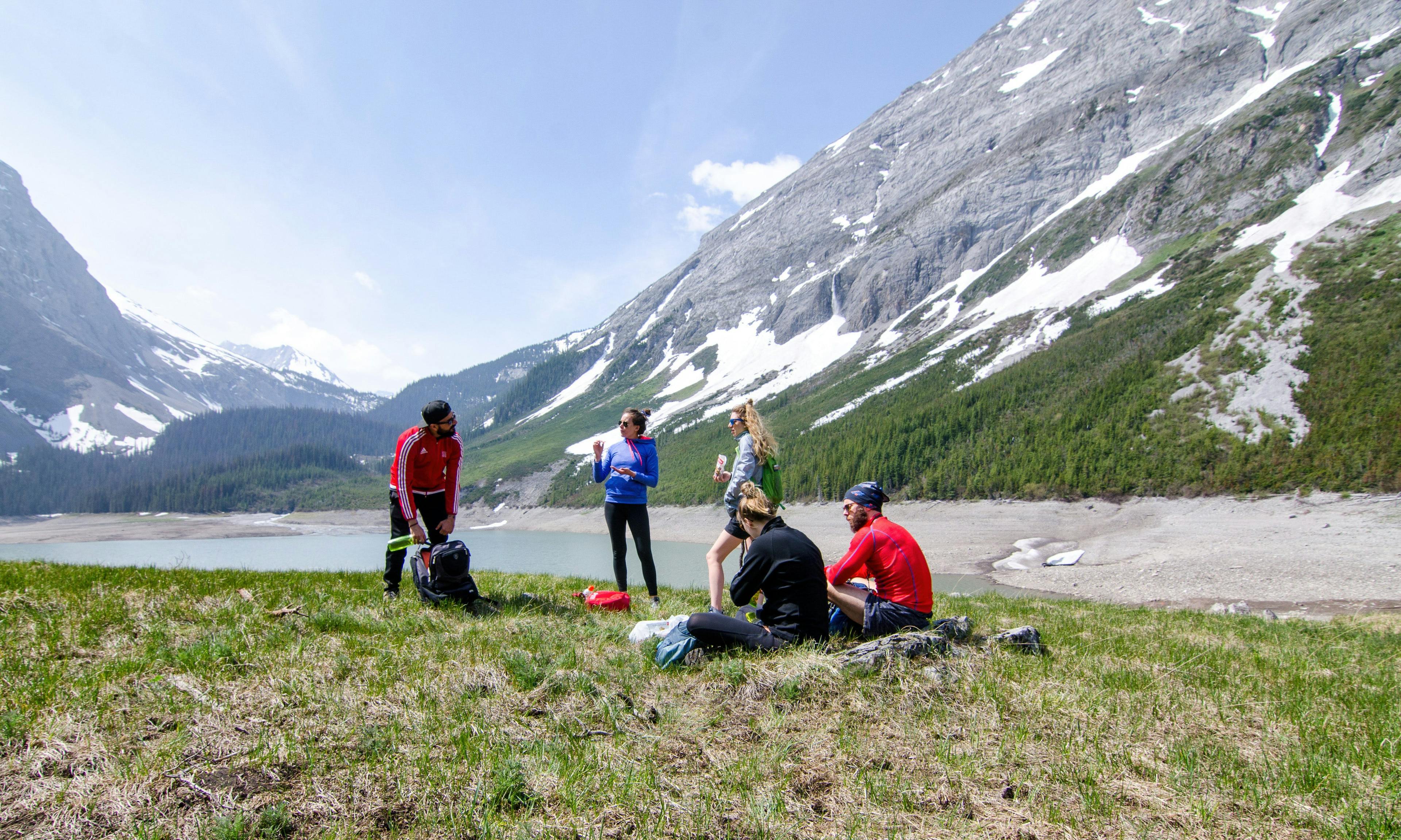 Five hikers eating lunch with large mountains and a lake behind them