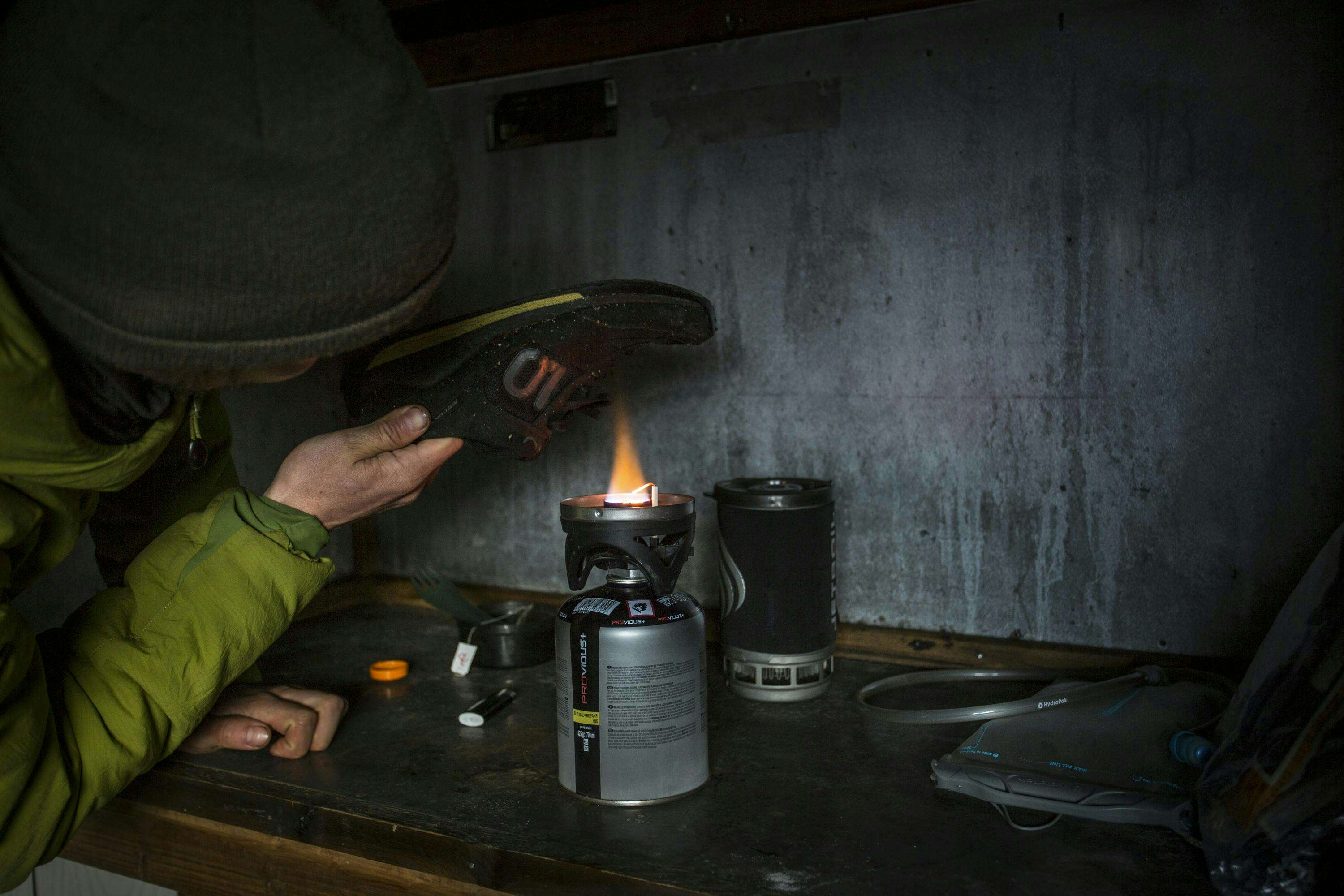 A hand holds a bike shoe over the open flame of a camp stove, trying to dry it out.