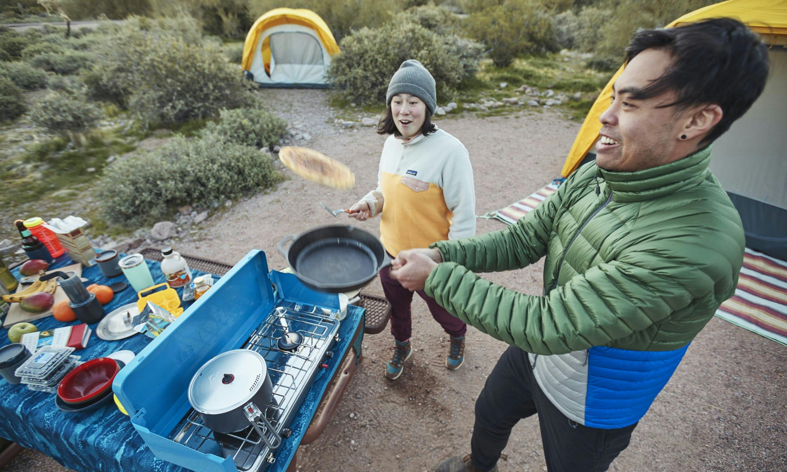 Camper flipping a pancake with tents and another camper in the background