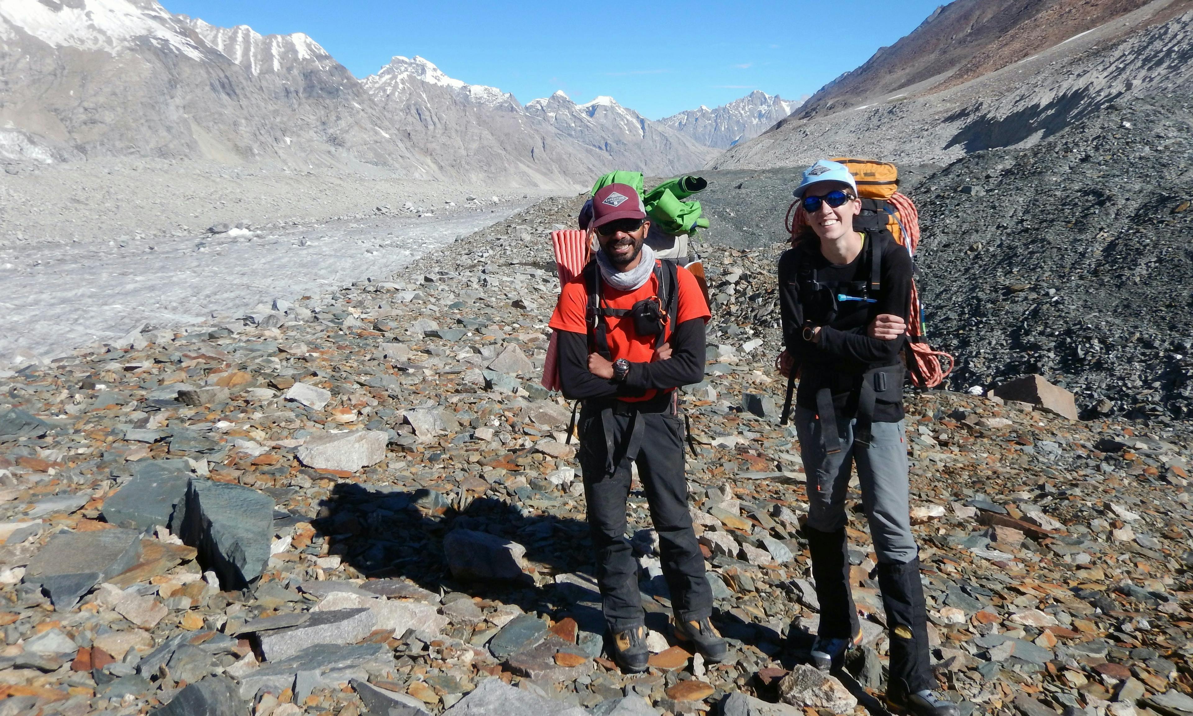 Two climbers wearing backpacks and standing on shale with mountains in the background.