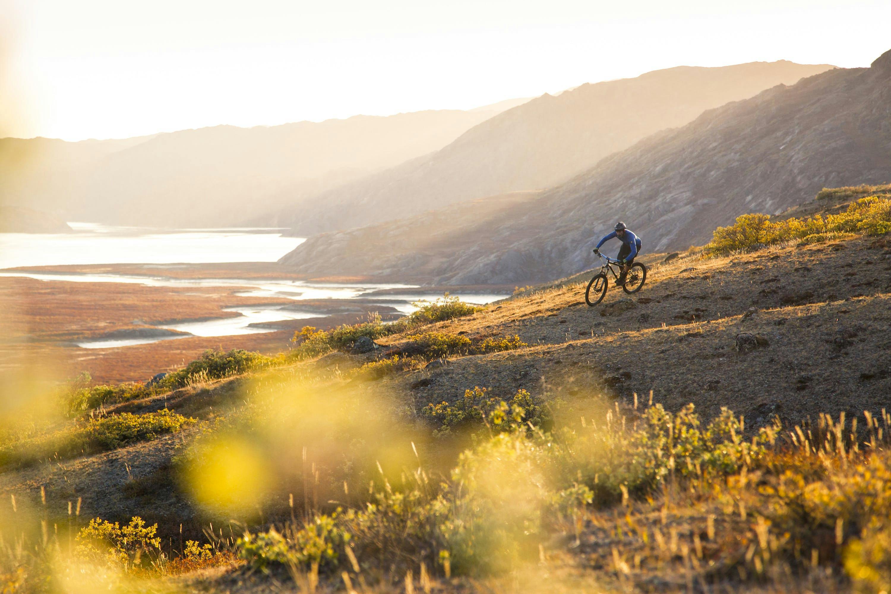 A lone cyclist rides across a grassy slope at golden hour with mountains behind him.