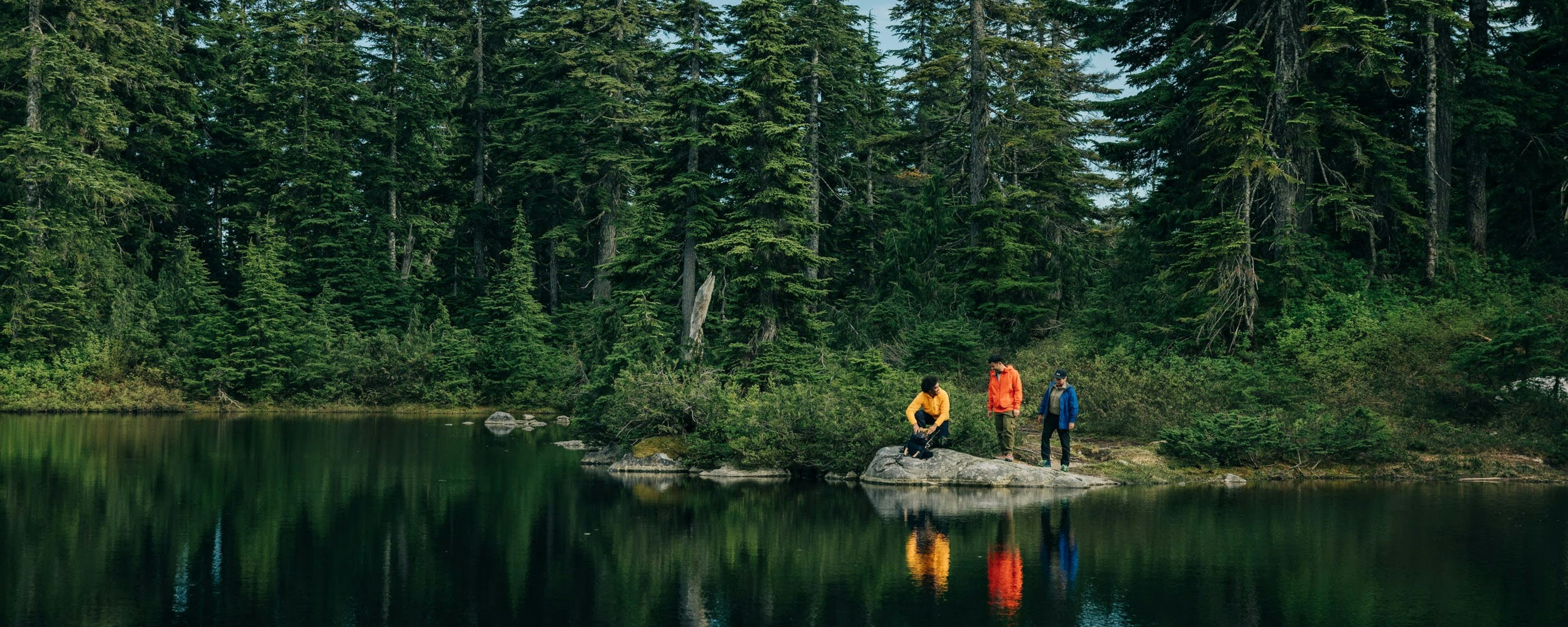 Three hikers stopping near the edge of an alpine lake with forest behind them