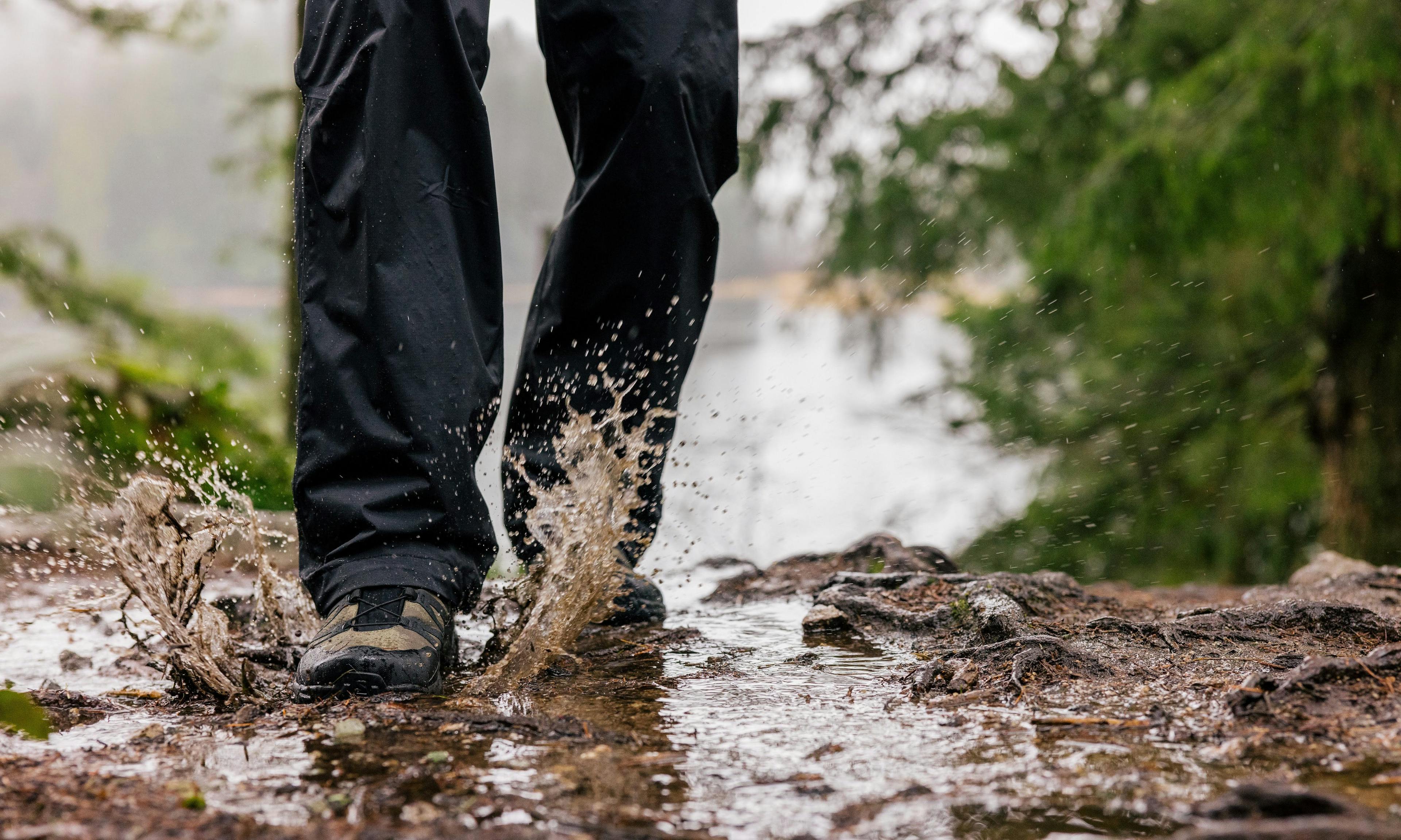 Randonneur en pantalon de pluie s'éclaboussant dans une grande flaque d'eau boueuse sur un sentier.