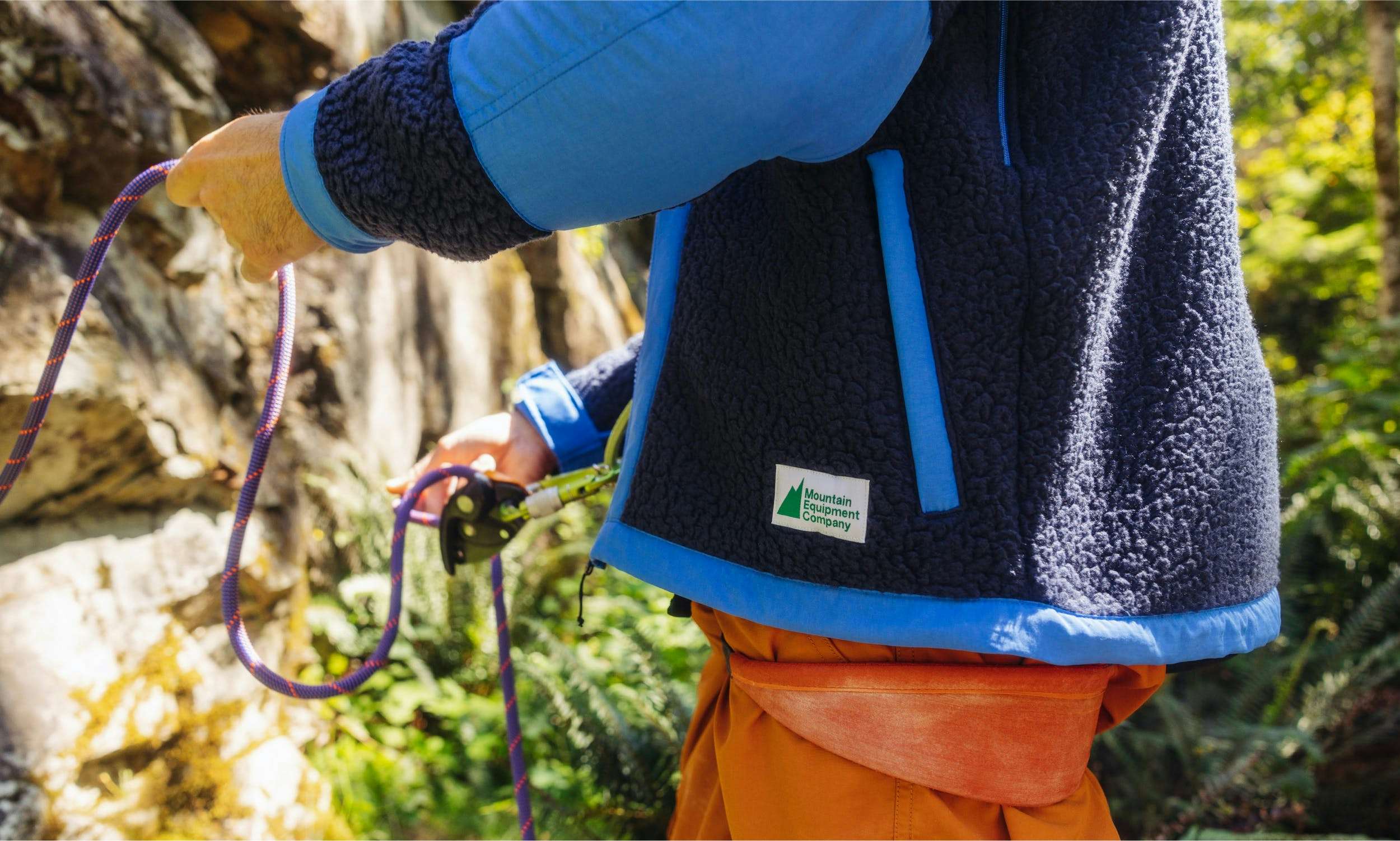 A closeup shot of a blue, fuzzy fleece with the MEC logo on the label in green. The person wearing the fleece is belaying.