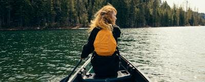 Women in the front of a canoe paddling on a lake. She is wearing a golden yellow PFD