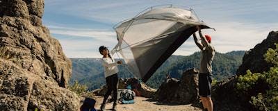 Two people lifting a setup tent above their heads. Campsite is in a rocky area